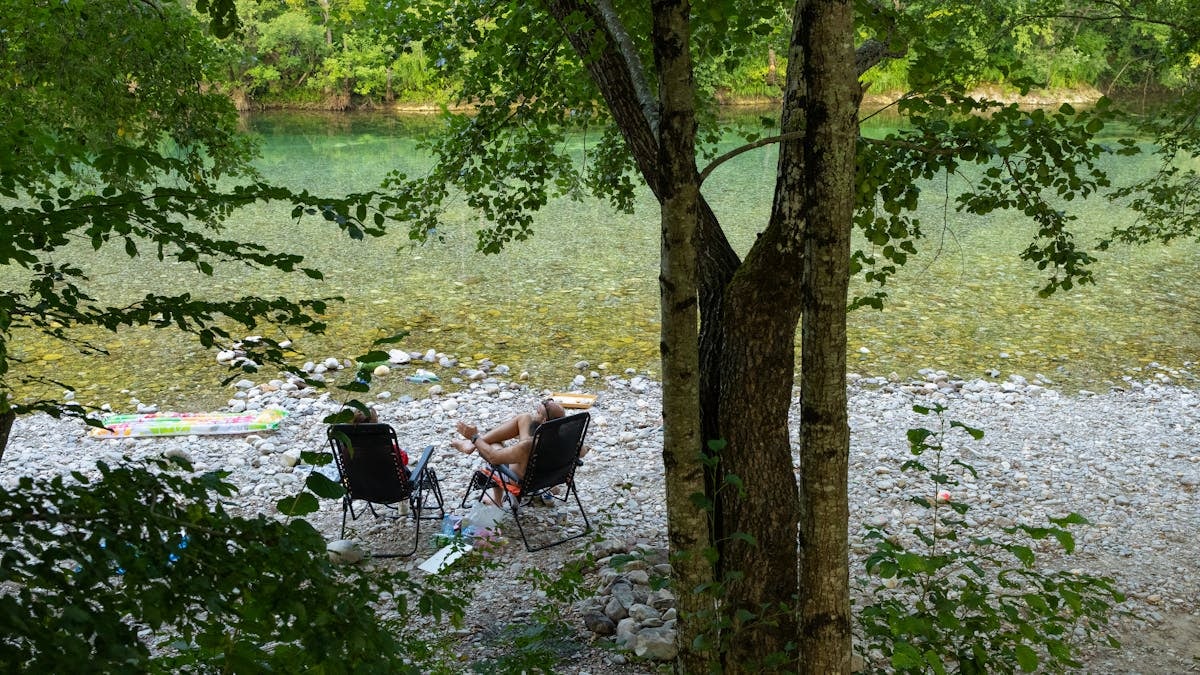 Two people relaxing on chairs by a clear riverbank surrounded by lush greenery.